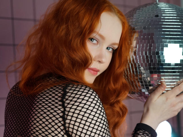 CheryCake, com cabelo ruivo ondulado, vestindo uma blusa preta de rede de pesca, segurando um espelho prateado brilhante, olhando por cima do ombro com um leve sorriso, mostrando sua pele clara..