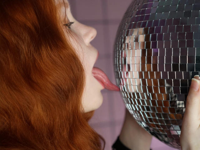 CheryCake, avec ses cheveux roux, la langue tirée, léchant une boule disco argentée brillante, la tenant avec sa main droite, montrant son reflet dans la surface miroir..