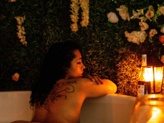 Scarlett, com cabelos pretos encaracolados, tatuagem nas costas, em uma banheira com pouca luz, sentada na borda, com velas acesas, decoração floral na parede, uma garrafa à direita..