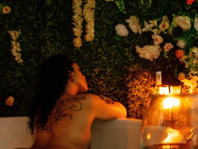 ScarlettBigAss, desnuda con cabello negro rizado, está sentada en una bañera, con un tatuaje en la espalda visible, mirando hacia arriba, con un farol encendido a la derecha y flores en el fondo..
