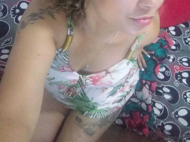 SolmaryHot with light skin, curly blonde hair, pink lipstick, floral dress, visible tattoos on arm and thigh, gold straps, necklace, looking down, red and black patterned background.
