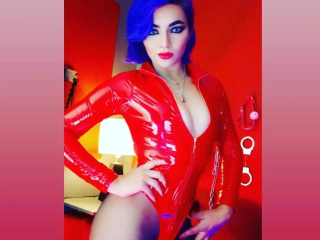MisssDarkLady met blauw haar, gekleed in een glanzend rood latex outfit met rits, waarbij haar borsten zichtbaar zijn, met haar hand op haar heup, rode lippenstift en een ketting, in een kamer met rood licht..