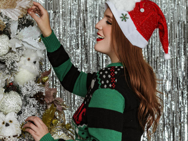 MelisaLee met lang bruin haar, een rode kerstmuts, een groen- en zwart gestreepte trui, die een witte kerstboom versiert, lachend, een tak vasthoudend, zilveren achtergrond, feestelijk, aantrekkelijk, borsten zichtbaar..