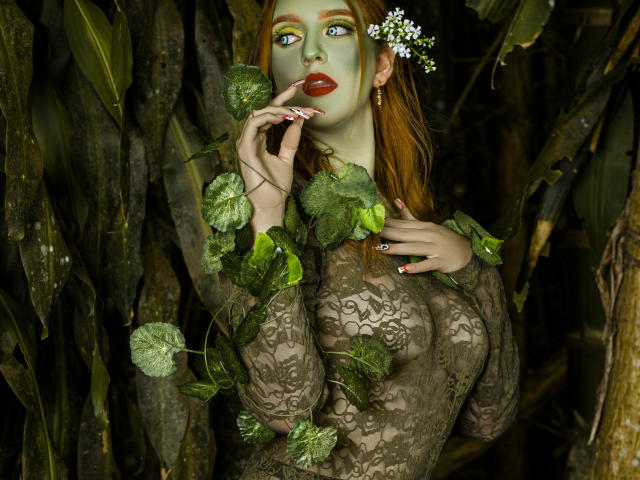 MelisaLee met groene make-up, rode lippen en een top van kant, omringd door bladeren, die haar vinger aan haar mond brengt, waarbij haar borsten zichtbaar zijn door de kant, met een witte bloem in haar haar..