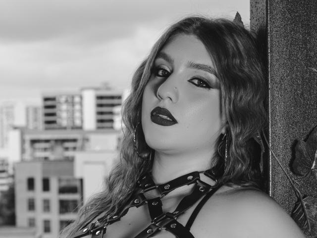 MelisaLee, com cabelo ondulado, batom escuro e uma coleira preta, apoiada em uma parede texturizada. Seios parcialmente visíveis, prédios da cidade borrados ao fundo. Foto em preto e branco..