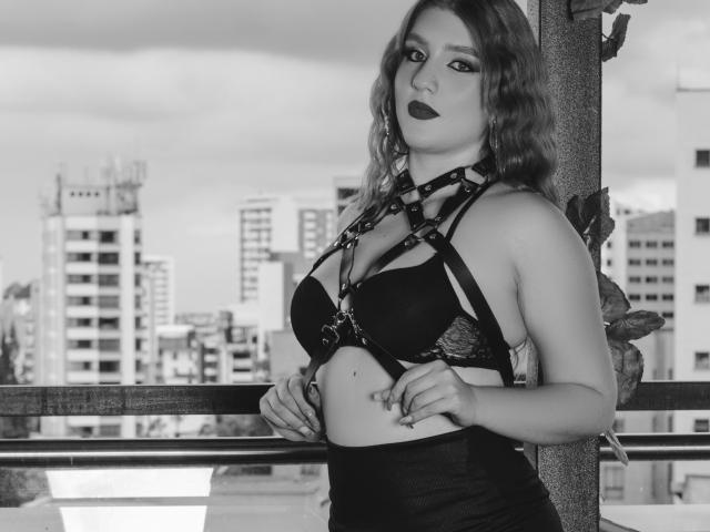Photo en noir et blanc de MelisaLee, avec des cheveux ondulés, du rouge à lèvres foncé, portant un soutien-gorge noir à bretelles fines, les mains sur sa taille, debout près d'une rambarde, avec des immeubles de la ville en arrière-plan..