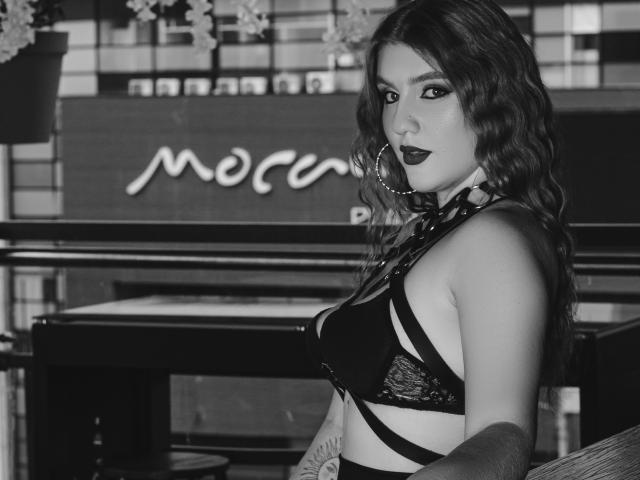 Black-and-white photo of MelisaLee with wavy hair, dark lipstick, large hoop earrings, strappy black lingerie, visible tattoo on right forearm, looking over her shoulder with a sultry expression.