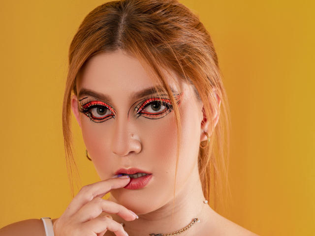 MelisaLee, com o dedo no lábio rosa, delineador vermelho e preto, cabelo loiro e brincos dourados, em um fundo amarelo, exibindo seu rosto adorável..