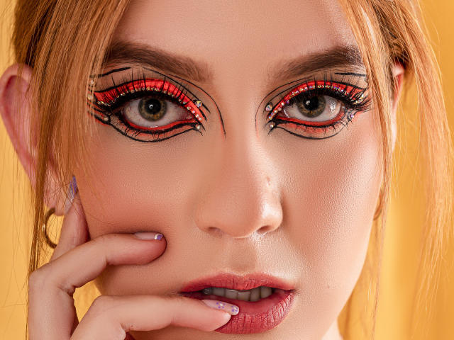 Het gezicht van MelisaLee, met rode en zwarte eyeliner, groene ogen en roze lippen, waarbij ze haar wang vasthoudt met bleekpaarse nagels, heeft een gele achtergrond en een lichte frons..