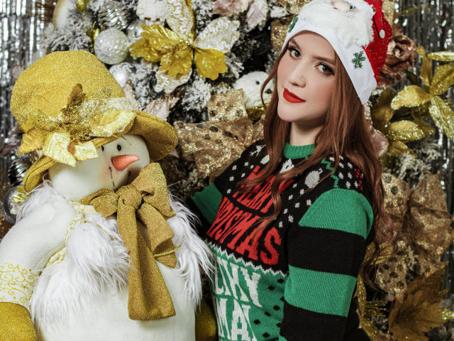 MelisaLee, com batom vermelho, usando um suéter listrado de verde e preto, e um chapéu de Papai Noel branco e vermelho, está em pé ao lado de um boneco de neve com um chapéu amarelo e um cachecol dourado. O boneco de neve tem um nariz laranja..
