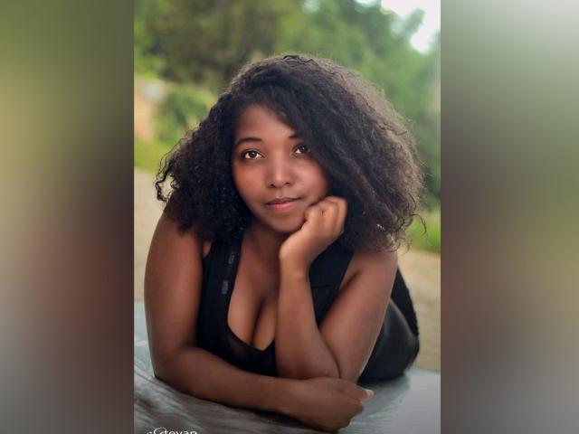 BlackCamilleX, com cabelo cacheado e escuro, pele morena e seios pequenos, vestindo uma blusa preta sem mangas, apoia a cabeça na mão, olhando para a câmera com um sorriso suave..