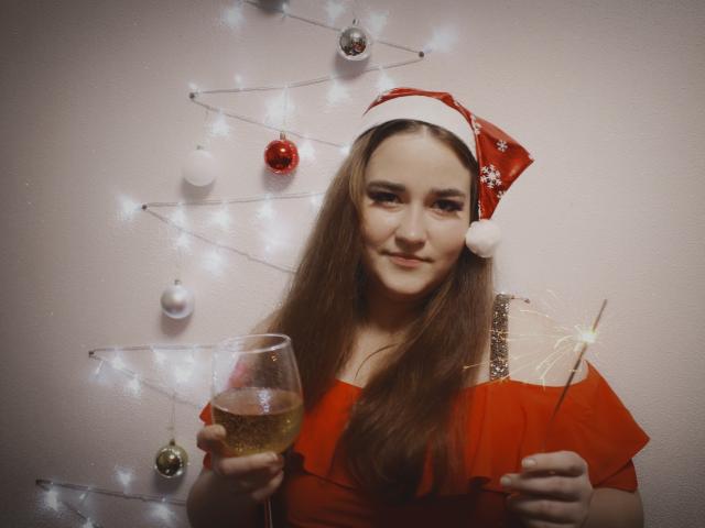DaniellaGraceZZ with long brown hair, wearing a red Santa hat and red dress, holding a glass of champagne and a sparkler, smiling. Christmas lights and ornaments in background.