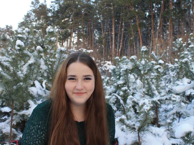 DaniellaGraceZZ with long brown hair, green knitted sweater, smiling slightly, standing in front of snow-covered pine trees. Her ass is not visible. She's cute.