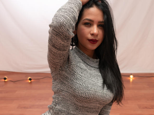 Stacy Kaplan, avec ses longs cheveux noirs, portant un pull gris en tricot, levant son bras, montrant ses seins, et regardant l'appareil photo avec du rouge à lèvres..
