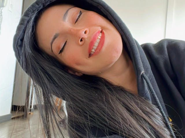 Liliysweet, con gli occhi chiusi e un sorriso sulle labbra, indossa un cappuccio nero, ha lunghi capelli neri e una felpa nera, mostrando un'espressione rilassata e felice. Il suo naso è leggermente cosparso di lentiggini..