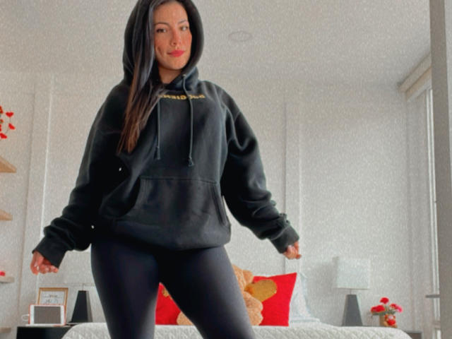 Liliysweet with long black hair, wearing a black hoodie and black leggings, standing in a room with a bed and red pillows, looking confident with a slight smile.
