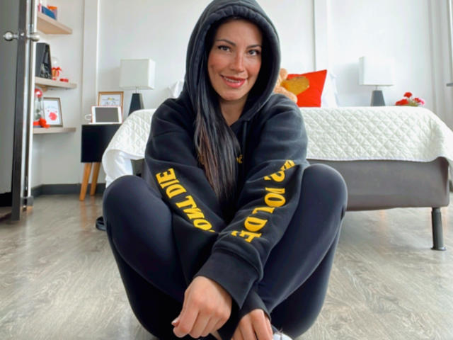 Liliysweet está agachada con las piernas cruzadas, vestida con una sudadera negra y pantalones negros, sonriendo a la cámara, con cabello largo y oscuro y un rostro lindo..