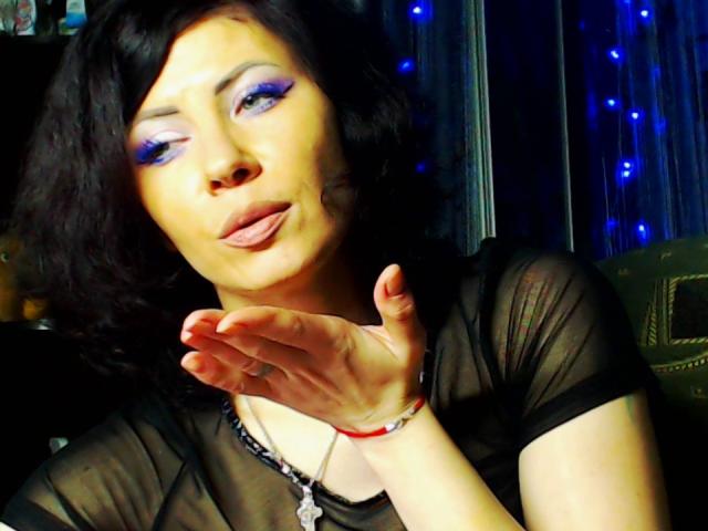 JuliaIce con sombra de ojos azul, cabello negro ondulado y una blusa negra transparente, haciendo pucheros con la mano cerca de su rostro, llevando un collar de plata y una pulsera roja..