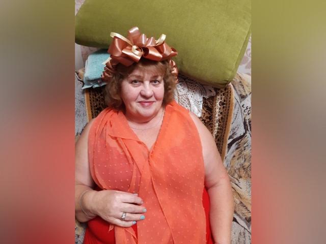 HelenAndChristy, een mollige vrouw met krullend bruin haar, gekleed in een glanzende oranje jurk en een grote bruine strik op haar hoofd, lacht naar de camera. Haar hand toont een ring..