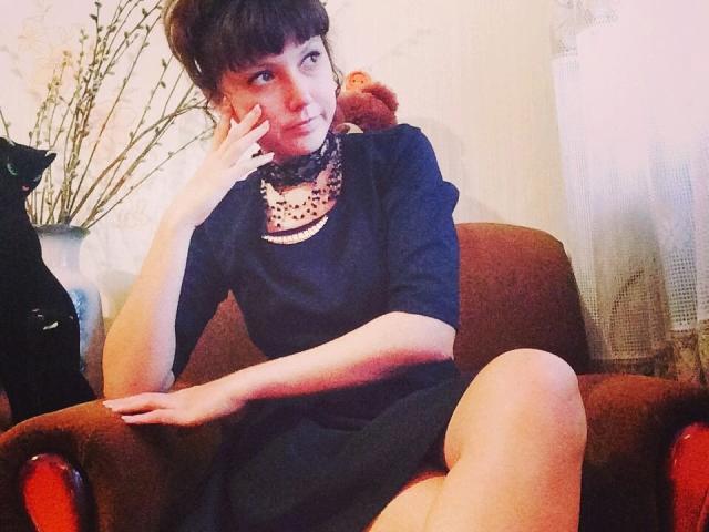 CarriePeachy, avec ses courts cheveux bruns, est assise sur un fauteuil marron, vêtue d'une robe noire et d'un collier, et elle touche sa joue, avec un ours en peluche marron derrière elle..