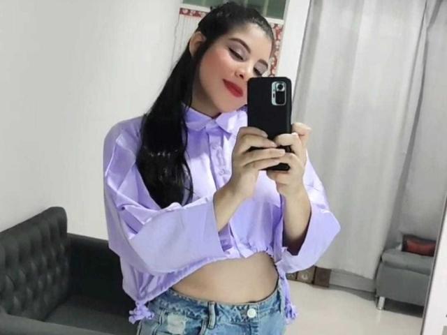 Charlote Evans, avec de longs cheveux noirs, du rouge à lèvres, portant une chemise lilas nouée au-dessus de son ventre, tenant un téléphone, prenant un selfie devant un miroir, portant un jean bleu délavé, souriante..