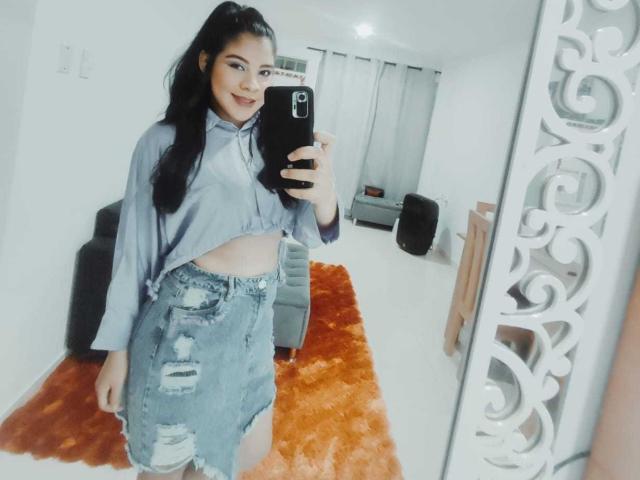 Charlote Evans, avec de longs cheveux noirs, souriante, portant un haut gris ample, une jupe en jean déchirée, et montrant son ventre plat, prend un selfie devant un miroir dans une pièce blanche..