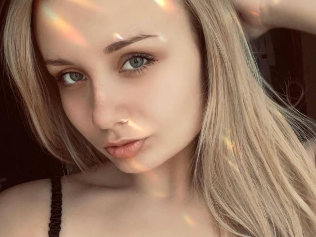 CherylGalaxy tem cabelo loiro claro, olhos azuis e um tom de pele claro, com um nariz delicado e lábios cheios. Ela está vestindo uma alça preta e olhando para a câmera..