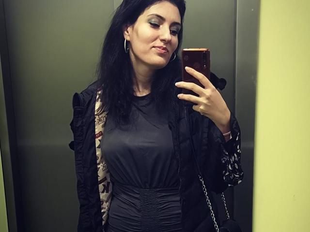 AddictedToUTS has dark hair, wearing a black dress with ruched waist, black jacket with floral sleeves, and a chain purse, taking a mirror selfie in an elevator.