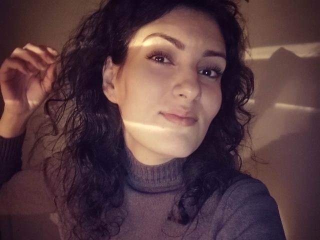AddictedToUTS with curly black hair, wearing a grey turtleneck, has light in her eyes, slight smile, and hand touching head. Soft shadows on face. Medium shot.