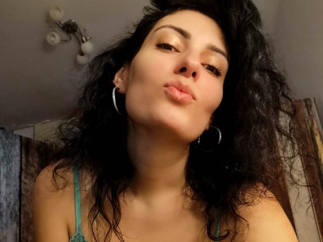 AddictedToUTS with curly black hair, hoop earrings, and a green top, pouting her lips, brown eyes, and light skin, in a low-light selfie, showing a slightly upturned nose.