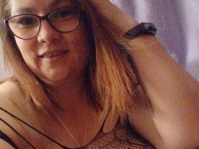 HellenBooty69 with glasses, smiling, wearing a black mesh top, showing her tits, arm raised, blonde hair, light skin, black wristband.