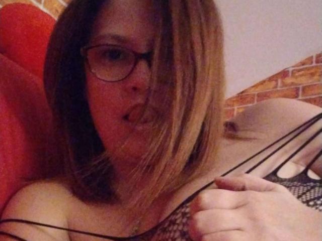 HellenBooty69 mit Brille, braunes, schulterlanges Haar, voller Lippen, hält ein schwarzes Netzoberteil, das kleine Brüste zeigt, bei schwachem Licht, leicht unscharf..