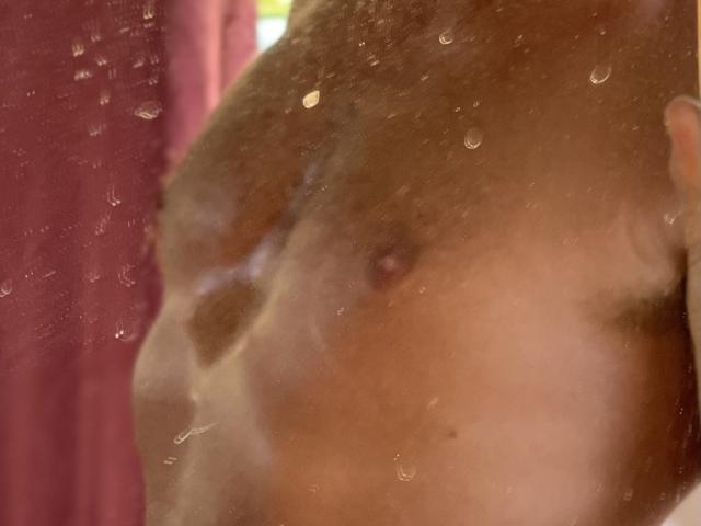 Die feuchte Brust von DomCaliente ist durch eine trübe, mit Wasser bedeckte Scheibe zu sehen, wobei kleine, braune Brustwarzen sichtbar sind. Wassertropfen sind über die Scheibe verteilt und zeigen Teile der Brust und des Oberkörpers..