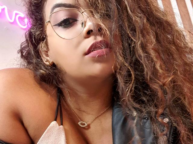 EbonyRose69, avec des cheveux bruns bouclés, des lunettes dorées, un eye-liner rose, du rouge à lèvres rose, un collier, du cuir noir et une serviette blanche, regardant vers le bas avec une expression légèrement boudeuse..