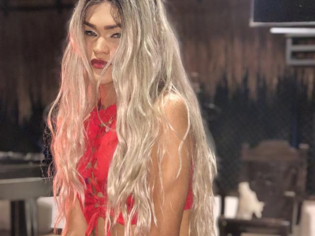 BarbTsX met lang, golvend, platina blond haar, draagt een rode, veter top zonder mouwen, waardoor haar borsten zichtbaar zijn. Ze heeft rode lippenstift en een serieuze, verleidelijke uitdrukking in een slecht verlichte kamer..