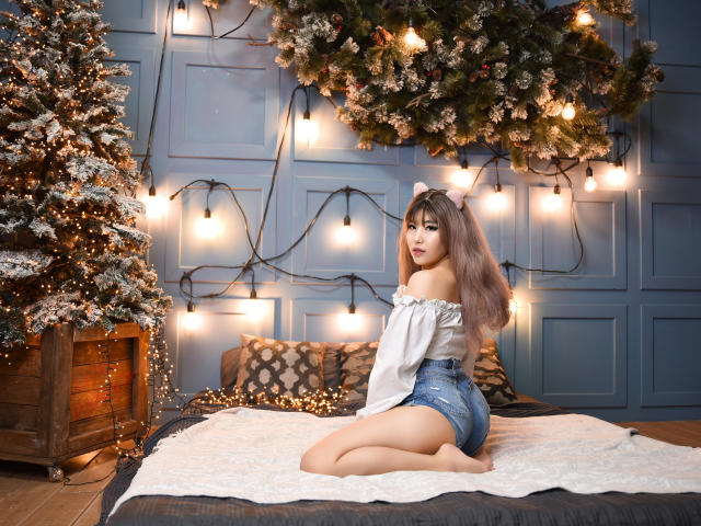 WendyFire, con el pelo largo y castaño y orejas de gato rosas, está sentada sobre una manta blanca, vistiendo una blusa con un hombro descubierto y pantalones cortos vaqueros, mostrando su trasero, al lado de un árbol de Navidad..