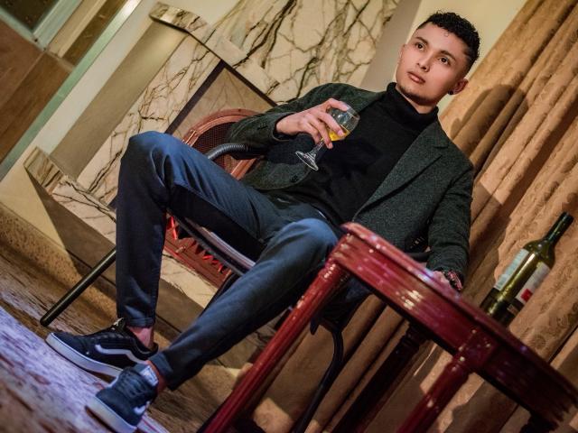 MelanyEtThomi, con cabello corto y oscuro, está sentada en una silla, sosteniendo un vaso de whisky, vistiendo un cuello alto oscuro, un blazer gris y jeans azules, con zapatillas negras, cerca de una mesa..
