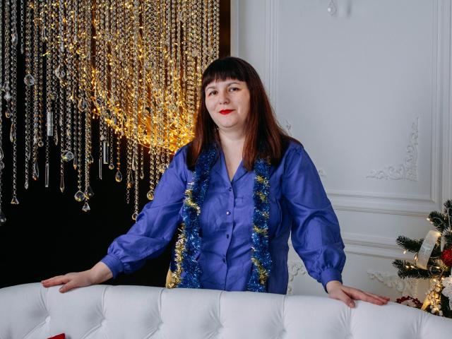 AliceInks with brown hair, wearing a blue satin shirt and blue/gold tinsel, standing with hands on white tufted sofa, red lipstick, looking at camera.