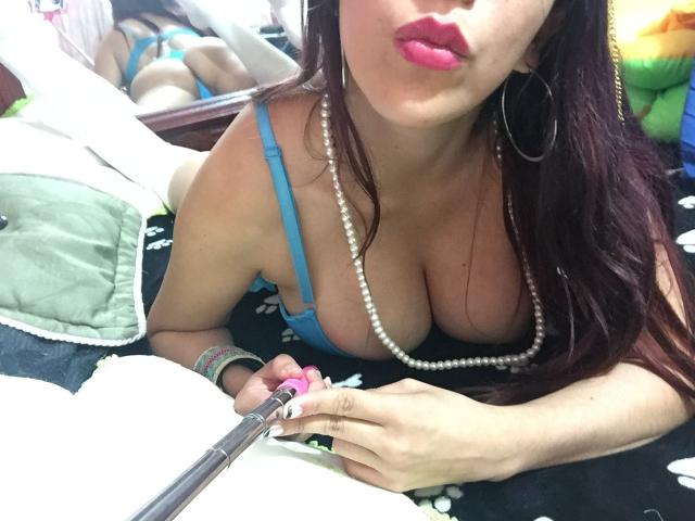 HotPamelaSex with big tits, wearing blue bra, pearl necklace, pink lipstick, holding pen, lying on bed, reflection of her ass in mirror, long brown hair, hoop earrings.