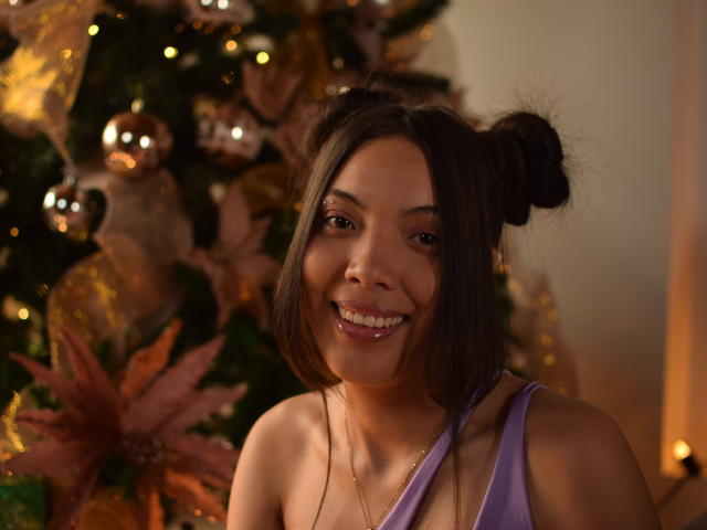ZharickTina met donkerbruin haar in twee knotjes, lachend, gekleed in een paars tanktop, waarbij haar borsten zichtbaar zijn, voor een versierde kerstboom, met warme verlichting, schattig..