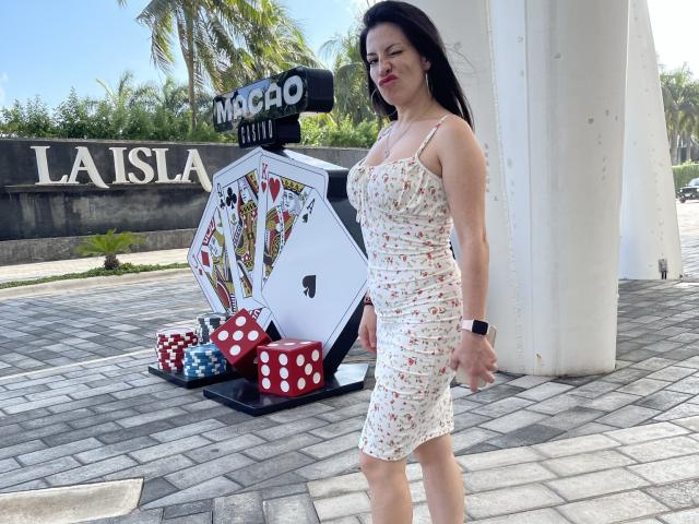 Liliysweet stands in a floral dress, showing her ass in a poker game setup with dice and cards, wearing a black wristwatch, smiling, black hair, sunny outdoor.