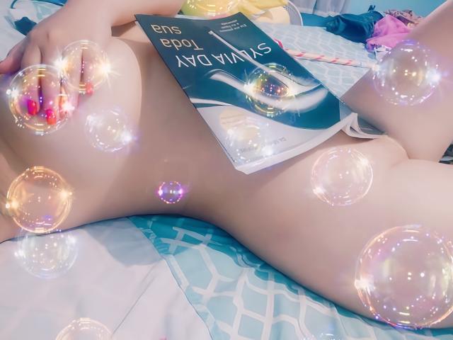 Il sedere pallido e nudo di AhriValvet è su un letto con un libro e un dildo rosa in mano, circondata da bolle scintillanti, sdraiata su un lenzuolo blu con motivi..