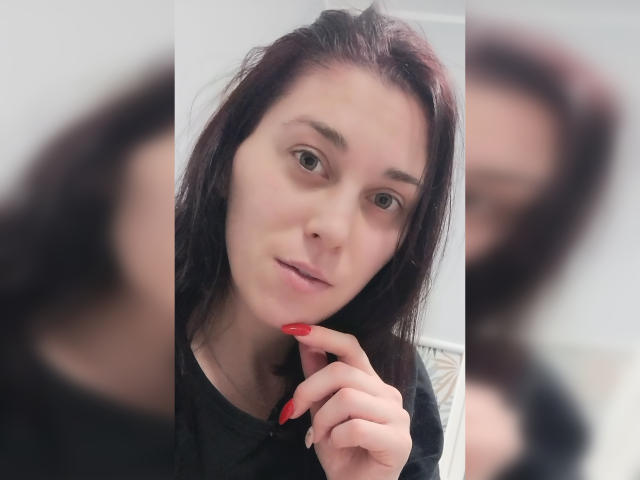BlackberryGil, com pele clara, olhos castanhos e cabelo ruivo escuro, vestindo uma camisa preta, tem unhas vermelhas e está tocando o queixo em uma selfie no espelho..