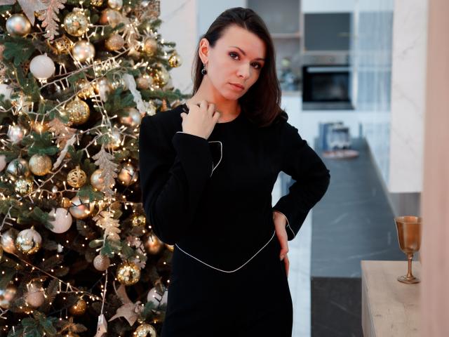 Sofia Foster está em um vestido preto, com a mão no pescoço, ao lado de uma árvore de Natal decorada com enfeites dourados e luzes. Ela tem cabelo castanho e uma expressão séria..