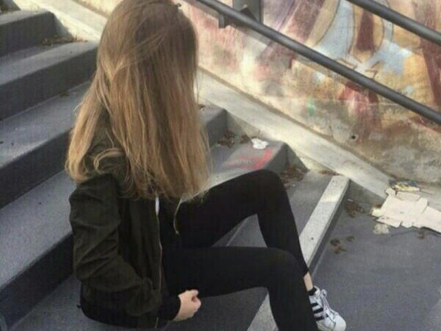 RoseJill, con cabello largo y castaño, chaqueta negra, pantalones negros y zapatillas blancas, sentada en unos escalones grises con la espalda hacia la cámara, con las manos sobre las rodillas..