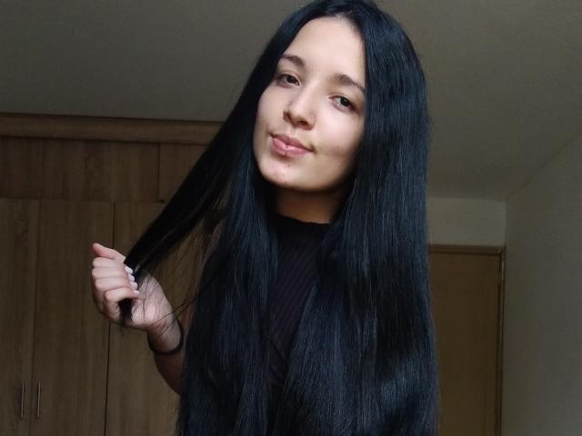 SarahGood with long black hair, holding a strand, smiling slightly, wearing a black shirt, brown eyes, light skin, in a simple room with wooden cabinets.