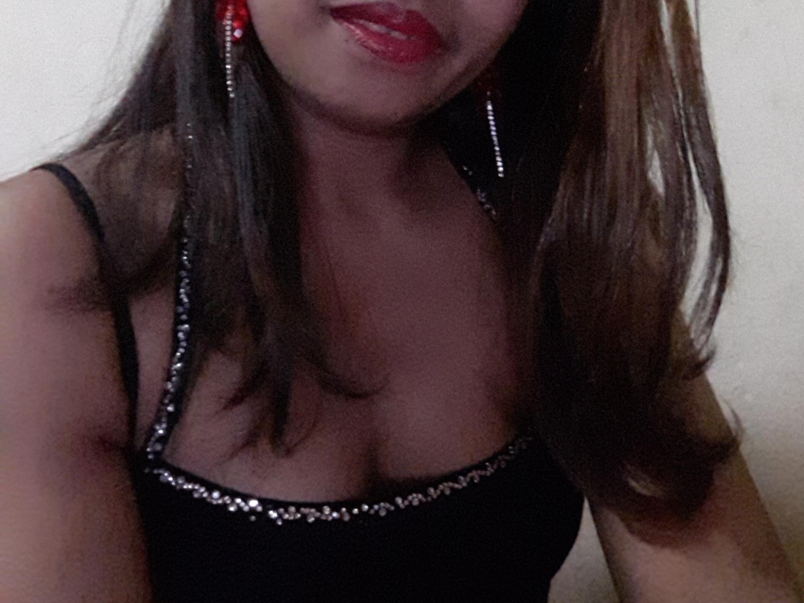 MayahLovely ha i capelli castani, le labbra rosse, un vestito nero con spalline e paillettes argentate, e orecchini rossi. In una foto scura e leggermente sfocata, mostra leggermente il seno..