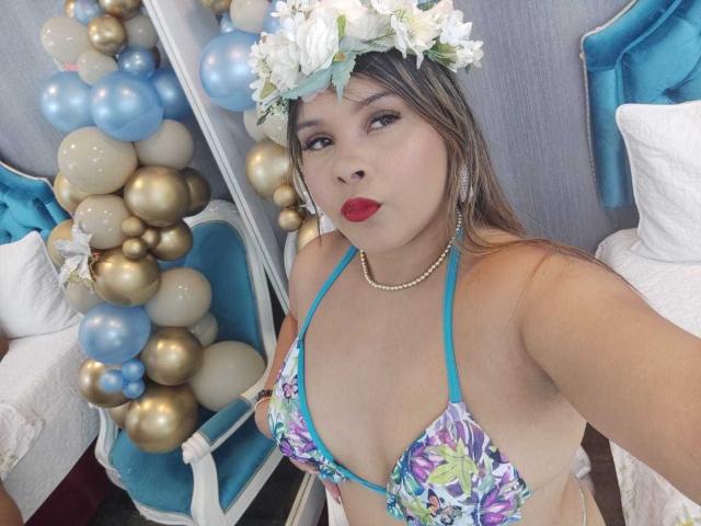 EvaMadison with a floral headpiece, wearing a blue and purple floral bikini top, showing her tits, red lipstick, pearl necklace, and blue balloons in the background.
