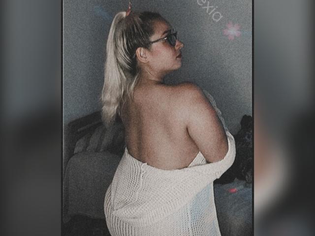 Alexia Morgan, com rabo de cavalo loiro, óculos e blusa branca de tricô com um ombro de fora, mostrando suas costas e nádegas, olhando por cima do ombro..