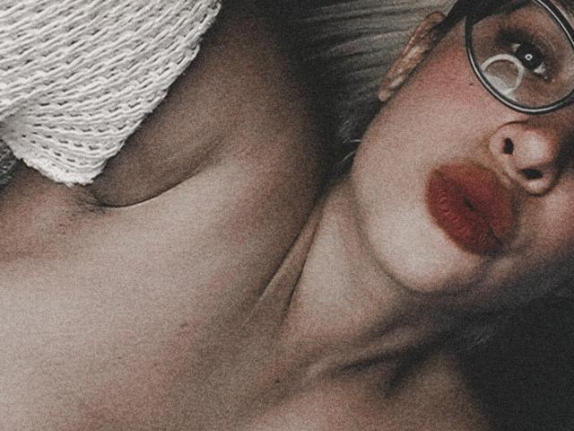 AlexiaMorgan con lápiz labial rojo, gafas y el brazo descubierto, acostada, vistiendo un suéter blanco de punto, mostrando su pecho desnudo y sus pequeños senos. Selfie granulada, con tonos oscuros..